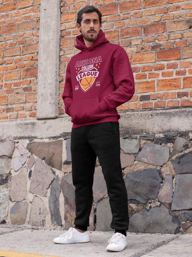 Man wearing a maroon hoodie with a logo, standing against a brick wall.