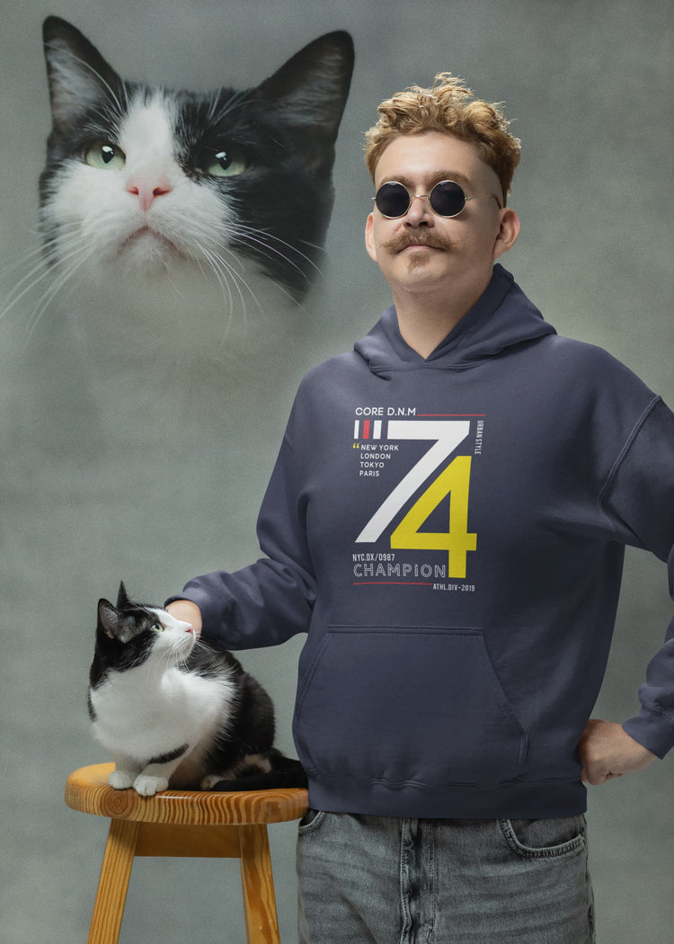 Man wearing a hoodie with a cat on a stool and another cat beside him against a gray background