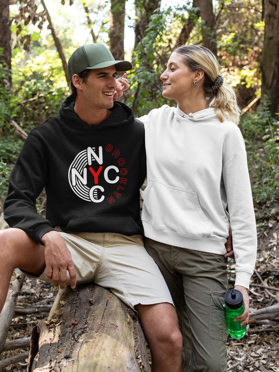 Two people sitting on a log in a forest, one wearing a black hoodie with a logo and the other in a white hoodie.
