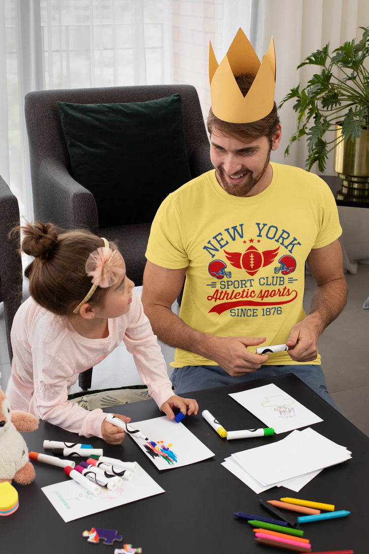 Man wearing a gold crown and yellow t-shirt with a child at a table with art supplies in a living room.