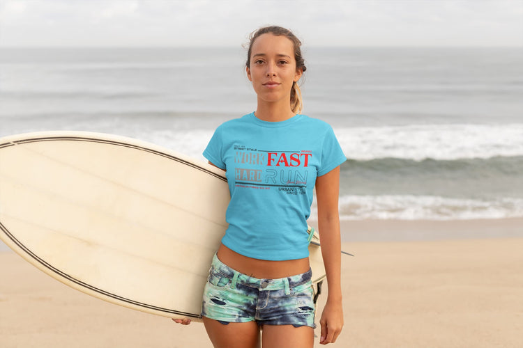 Person holding a surfboard on a beach wearing a blue t-shirt with text and patterned shorts.