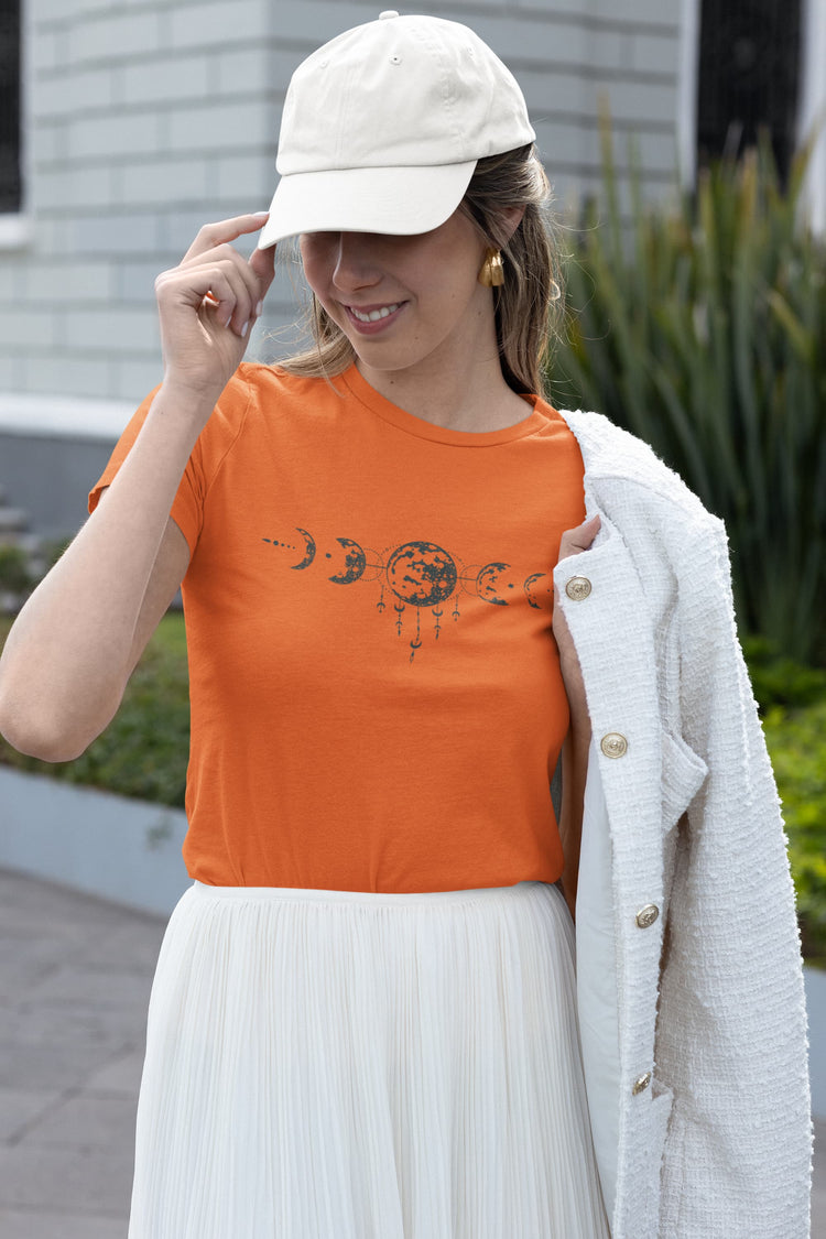Woman wearing an orange t-shirt with a graphic design, white skirt, and beige cap outdoors.