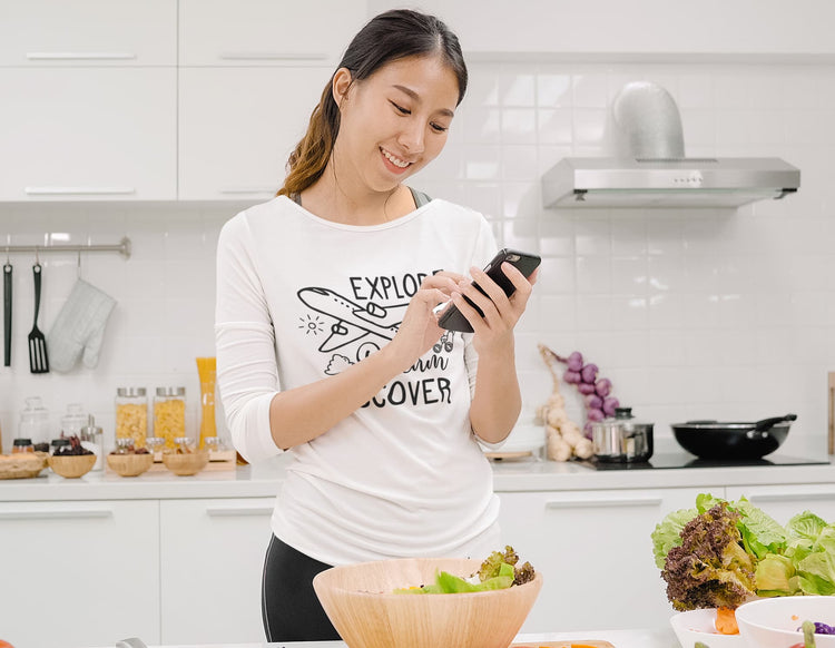 Woman using a smartphone in a kitchen with various items on the counter.