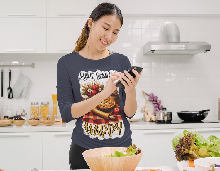 Woman in a kitchen wearing a shirt with a pie design and text, using a phone.