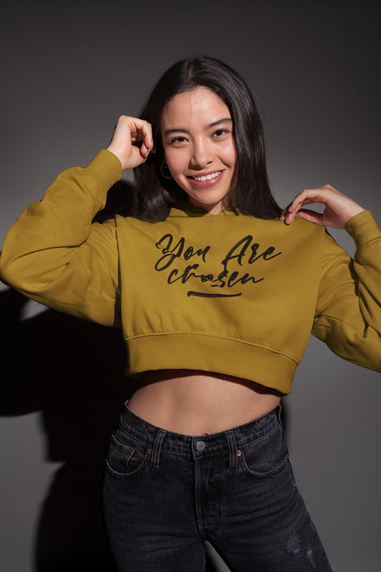 Woman wearing a mustard yellow crop top with text, posing against a dark background