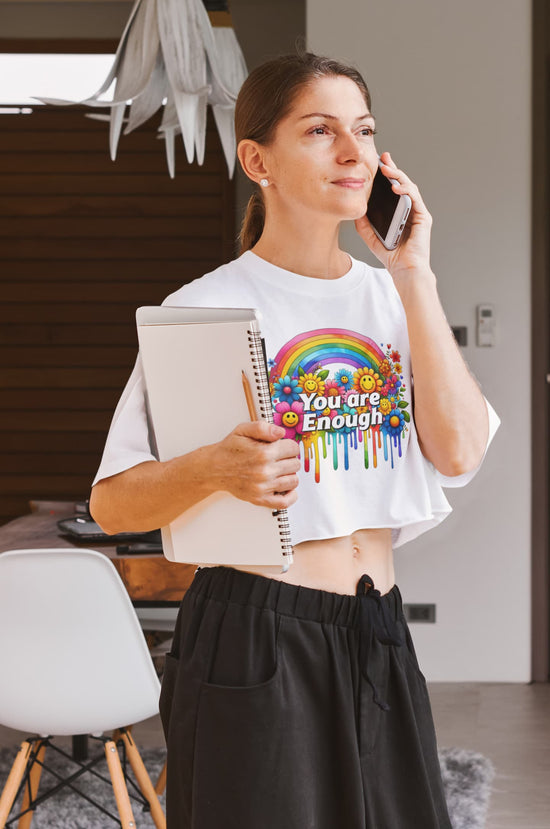 Woman holding a notebook and phone, wearing a t-shirt with a colorful design and text.