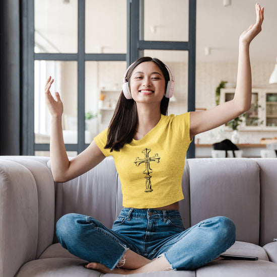 Woman sitting on a couch wearing headphones and a yellow shirt with a cross design, arms raised in a living room.