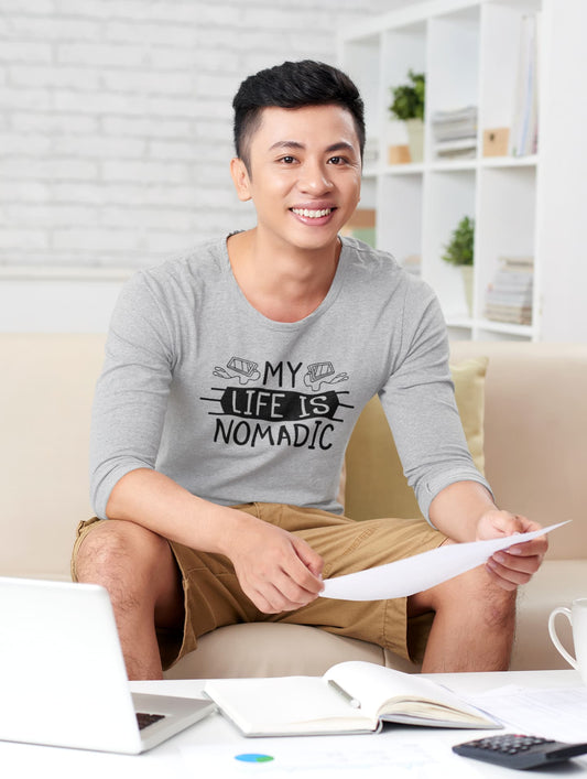 Man sitting on a couch with a laptop and papers, wearing a gray shirt with 'My Life is Nomadic' text.