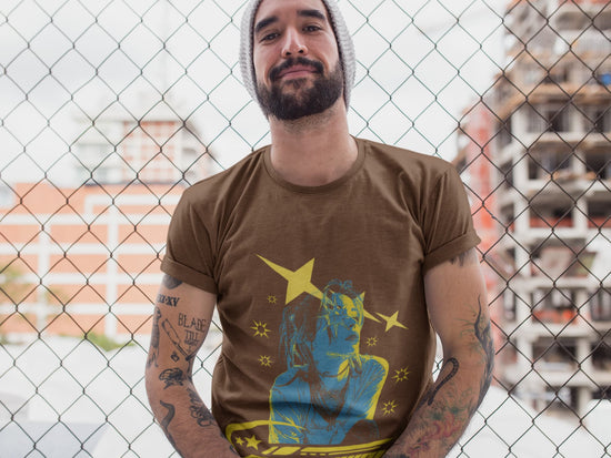 Man wearing a brown t-shirt with a graphic design in front of a chain-link fence.