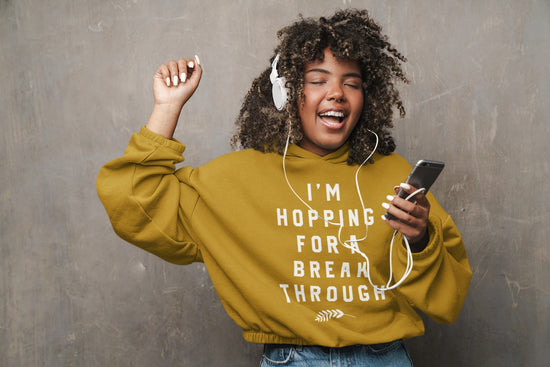 Person wearing a mustard yellow hoodie with text, listening to music on headphones against a gray background