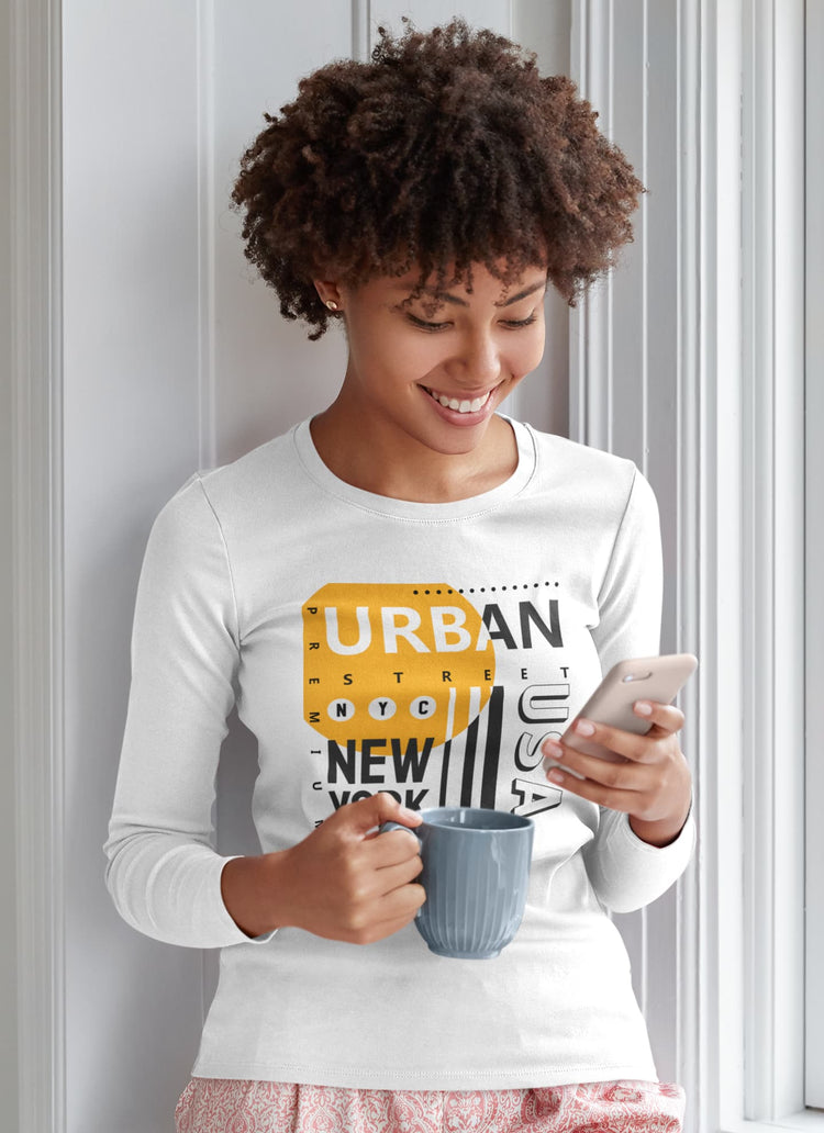 Woman wearing a white long-sleeve shirt with a graphic design, holding a mug and phone, standing against a light-colored wall.
