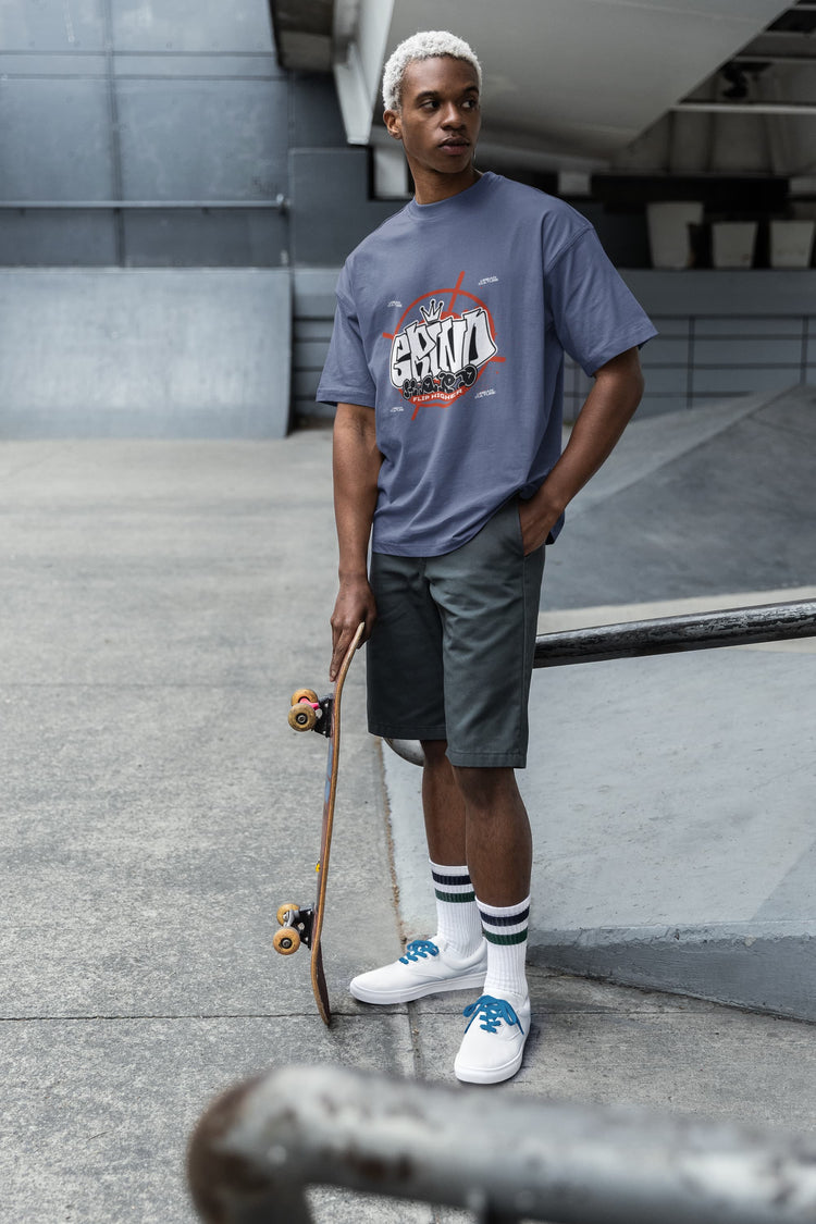 Man holding a skateboard wearing a gray t-shirt with a logo and text, standing on a concrete surface.
