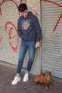 Person wearing a hoodie and jeans standing next to a dog on a street with graffiti.