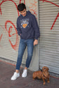 Person wearing a hoodie and jeans standing next to a dog on a street with graffiti.