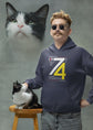 Man wearing a hoodie with a cat on a stool and another cat beside him against a gray background
