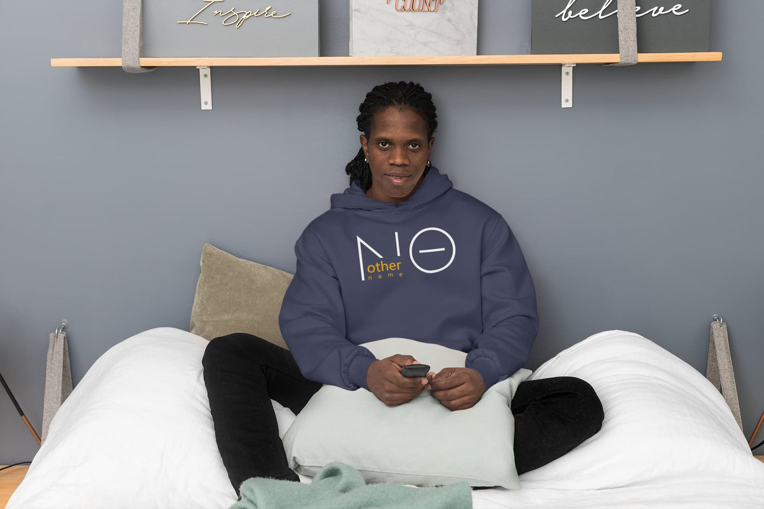 Person sitting on a bed wearing a hoodie with a logo, holding a phone, against a gray wall with shelves displaying motivational word pillows.