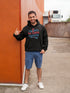 Man wearing a black hoodie with colorful text and blue shorts standing against an orange wall.