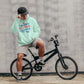 Person wearing a light green hoodie with text, sitting on a black bicycle against a gray wall.