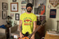 Person wearing a bright yellow t-shirt with text, holding a guitar in a room with various decorations.