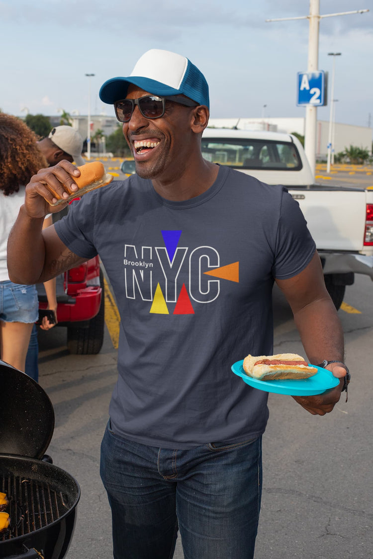 Man wearing an NYC t-shirt holding a hot dog and a plate with another hot dog, standing outdoors.