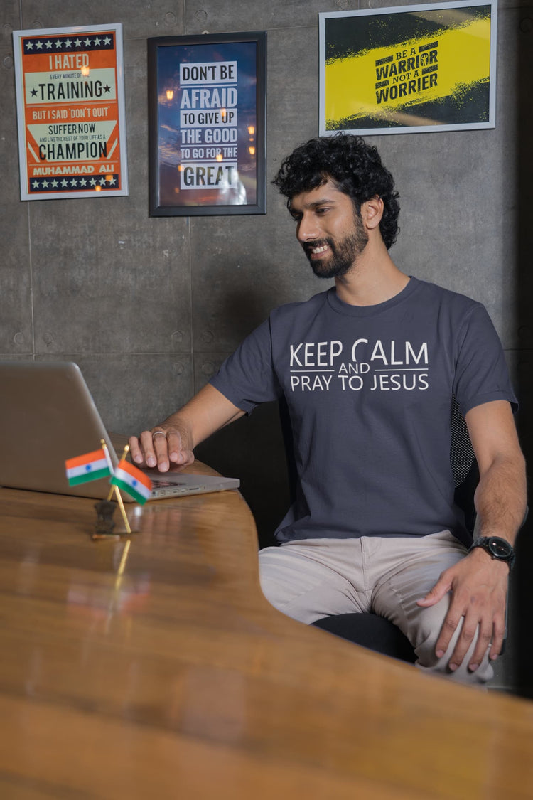 Man sitting at a desk with a laptop, wearing a t-shirt with a religious message, in an office setting.