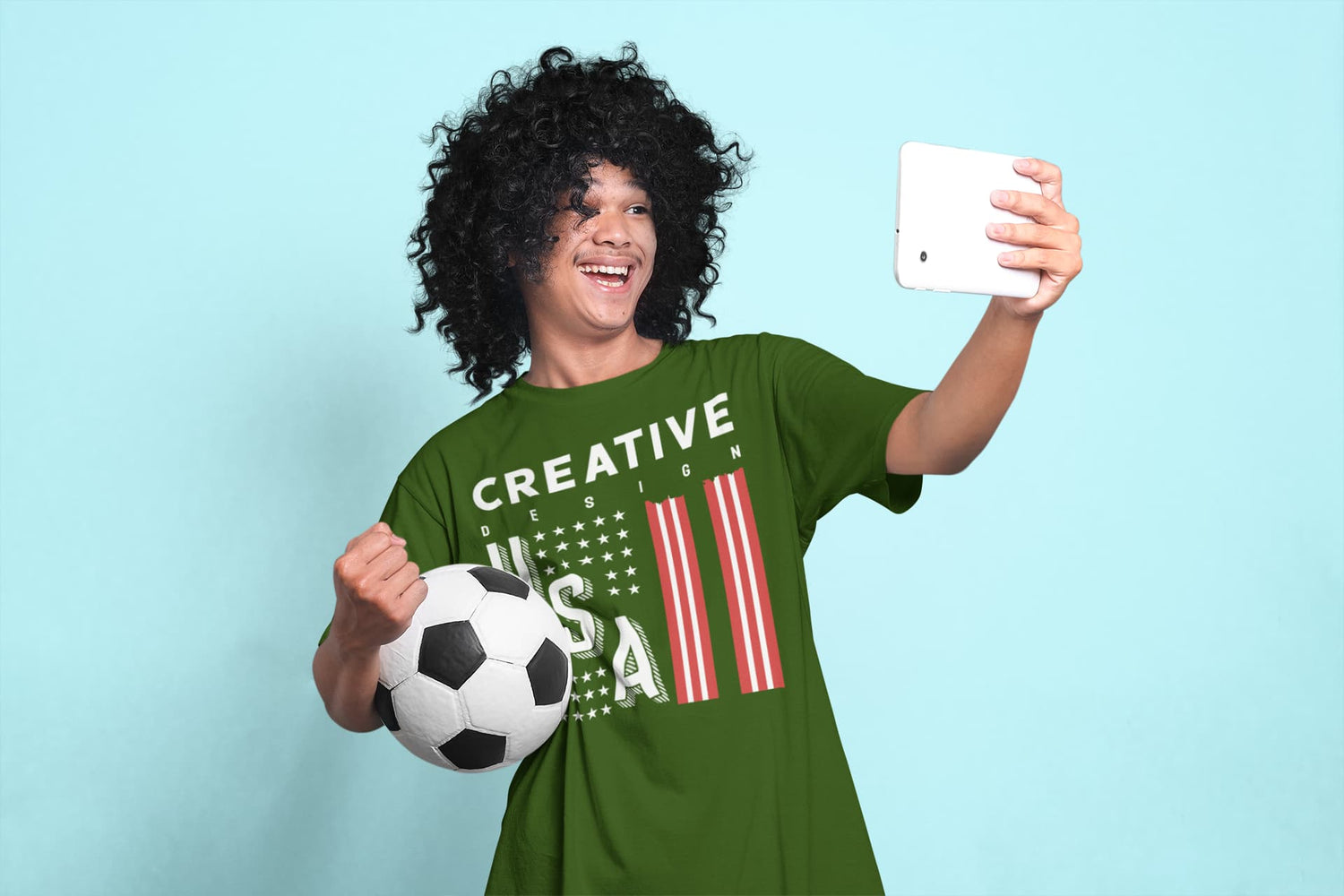 Person taking a selfie with a soccer ball wearing a green t-shirt with USA design on a light blue background