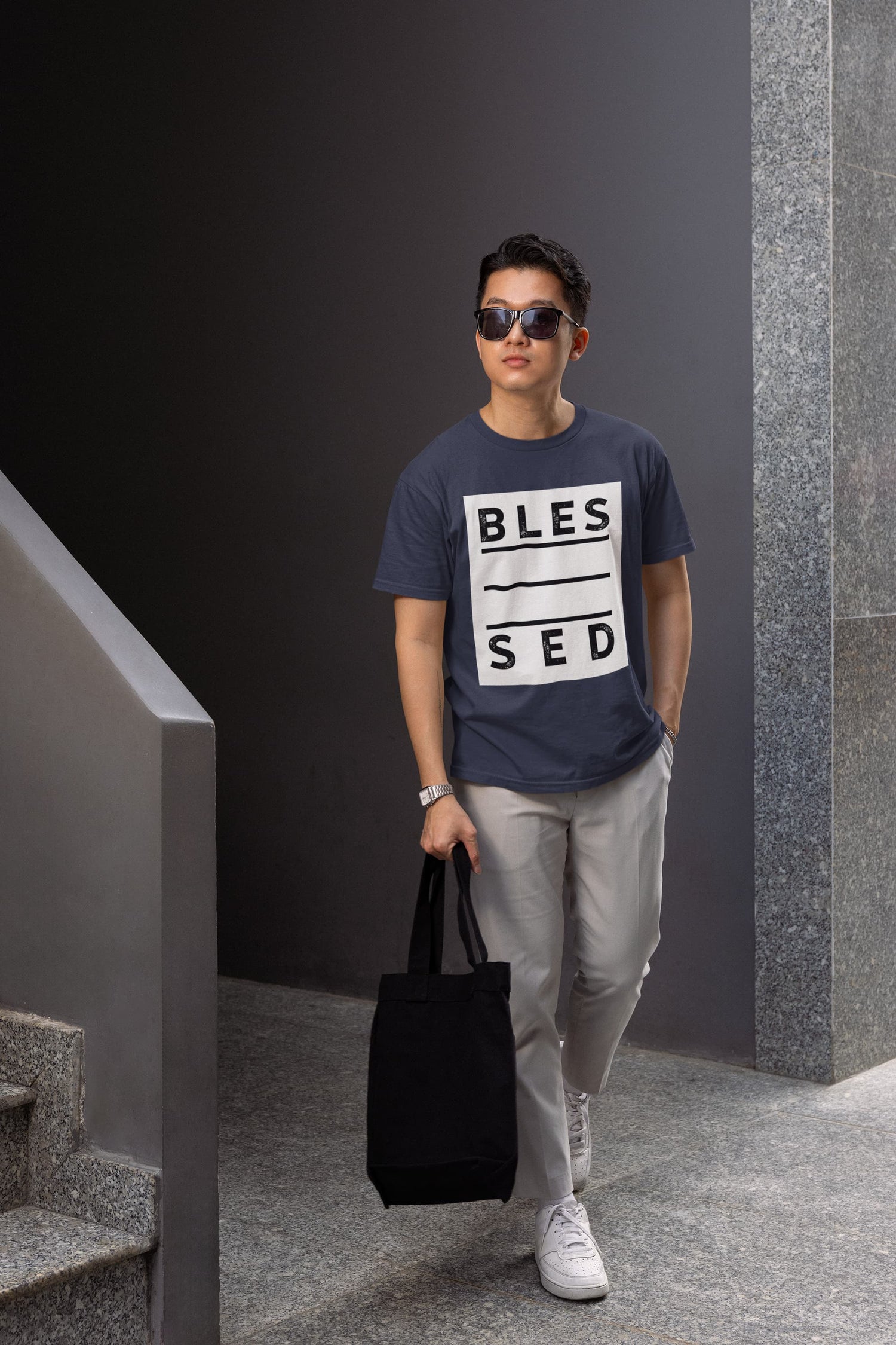 Man wearing a navy blue t-shirt with text, holding a black bag, standing in a modern indoor setting.