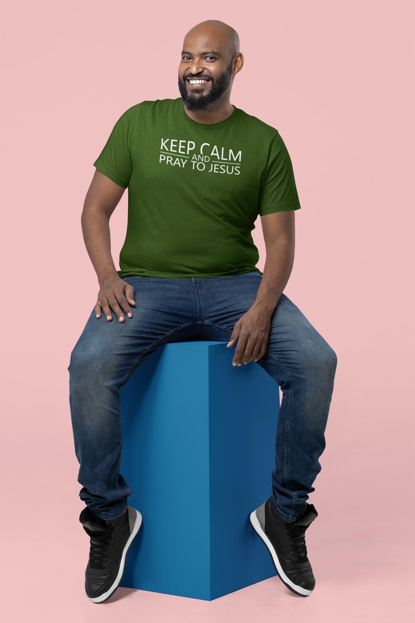 Man sitting on a blue stool wearing a green t-shirt with text, against a pink background