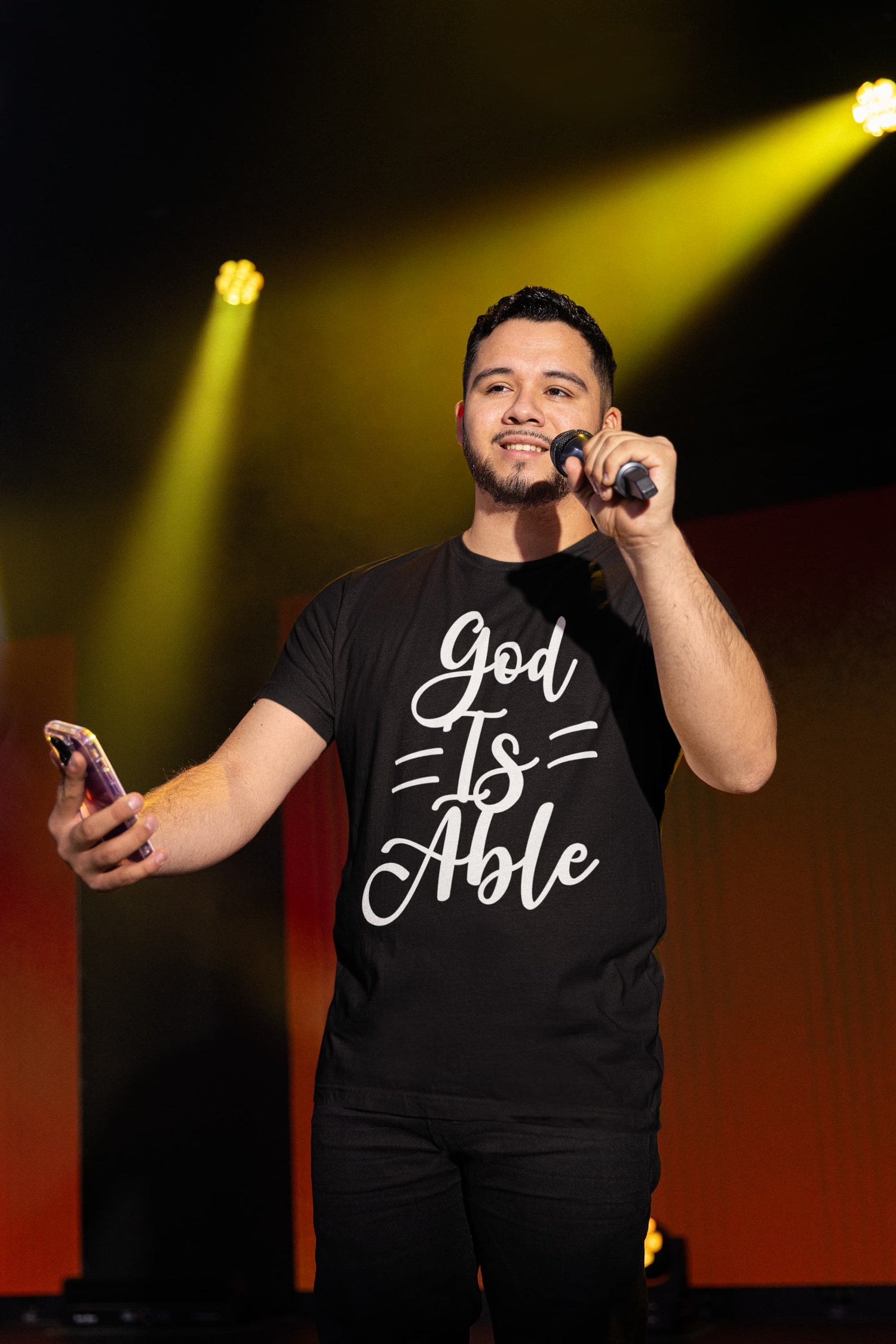 Man on stage holding a microphone and phone, wearing a black t-shirt with &