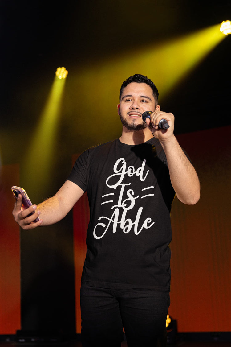 Man on stage holding a microphone and phone, wearing a black t-shirt with &