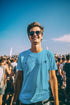 Man wearing sunglasses and a blue t-shirt at a sunny outdoor event.