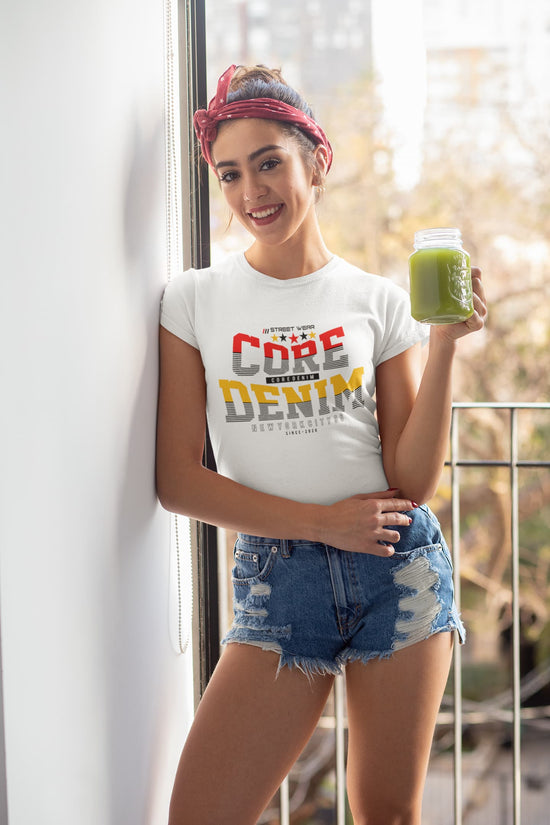 Woman wearing a white t-shirt with colorful text, holding a green smoothie, standing indoors.