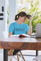 Woman in a blue t-shirt sitting at a desk with a laptop and book, holding a coffee mug.