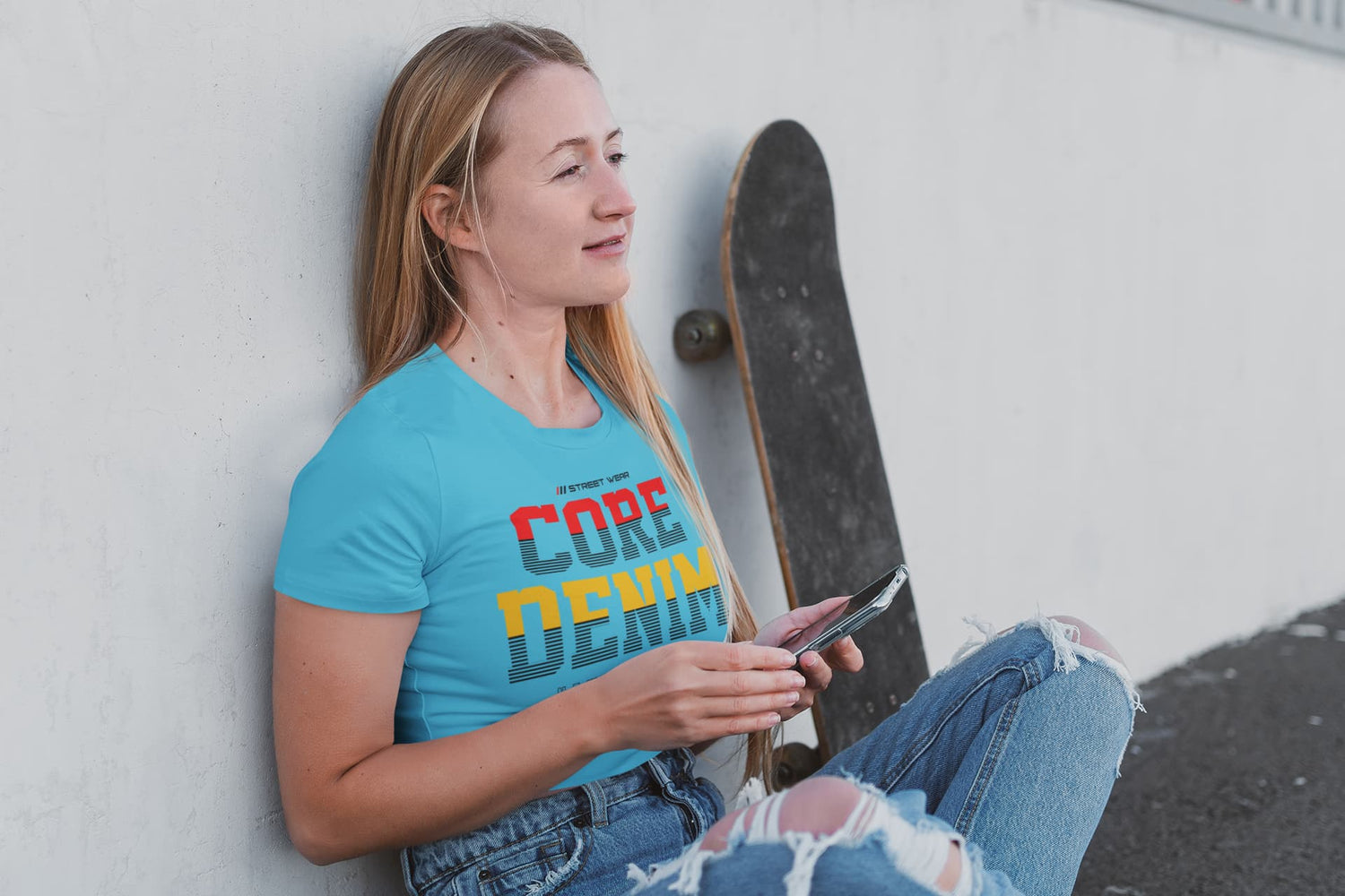 Woman wearing a blue t-shirt with colorful text, holding a skateboard and a phone, against a light-colored wall.