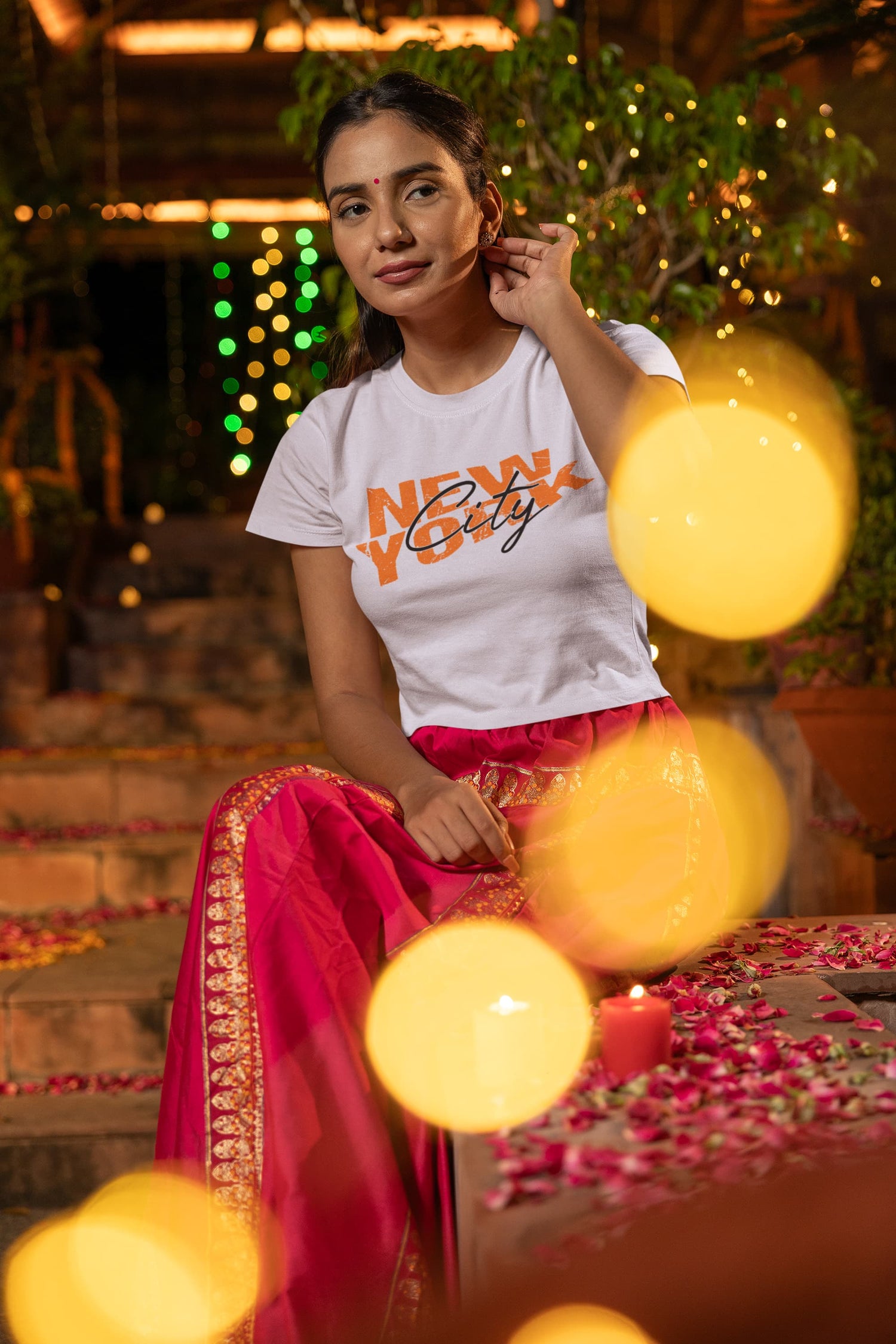 Woman in a white t-shirt with text and pink traditional outfit sitting outdoors with blurred lights in the background