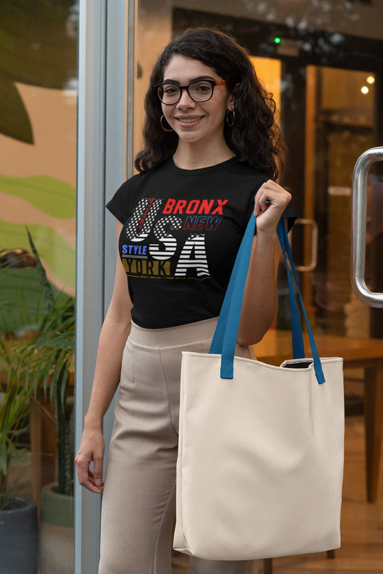 Woman holding a tote bag, wearing a black t-shirt with text, in an indoor setting.