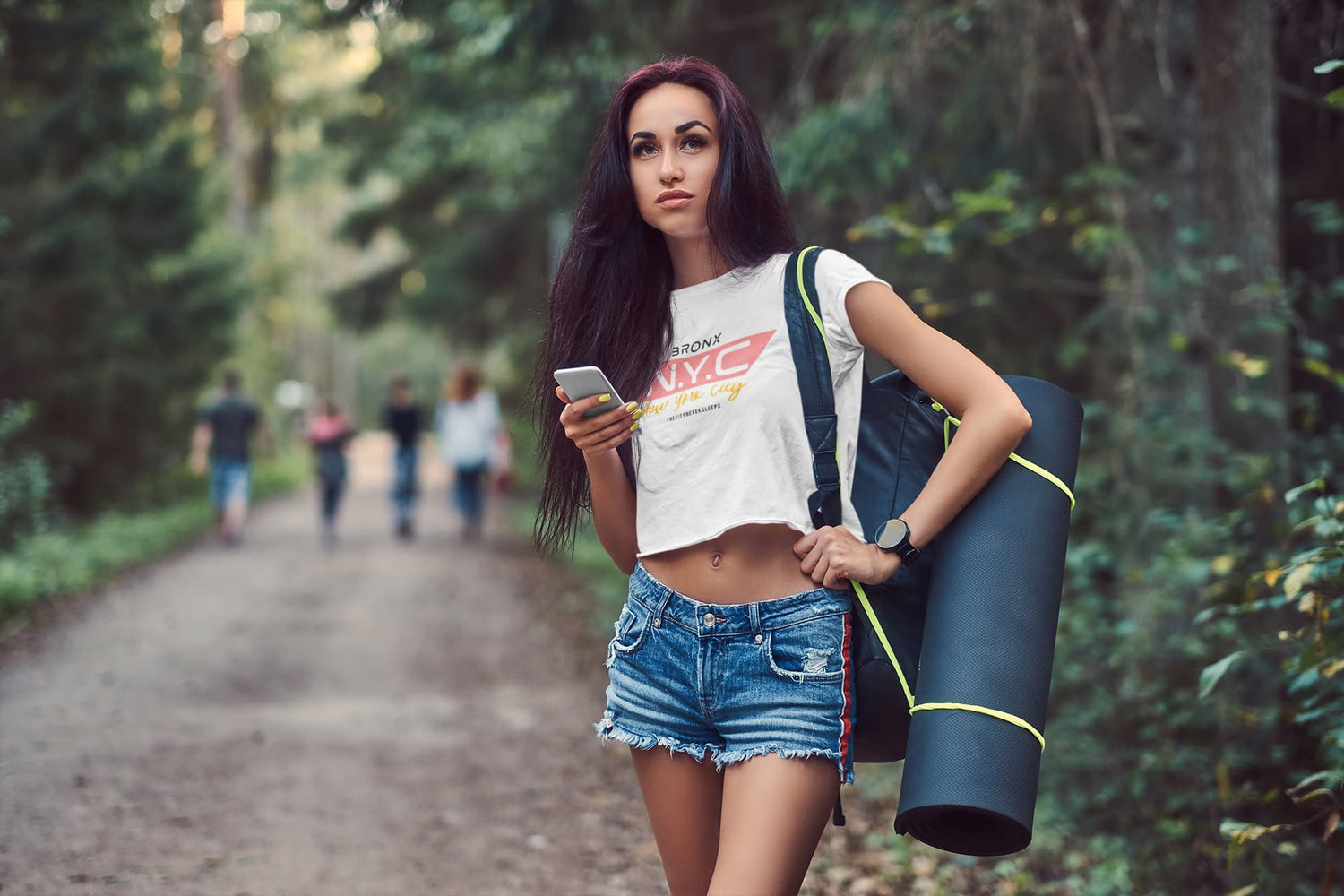 Woman walking in a forest holding a phone and a blue mat, with blurred people in the background.