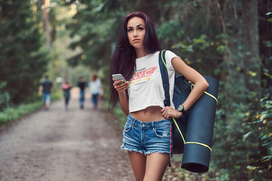 Woman walking in a forest holding a phone and a blue mat, with blurred people in the background.
