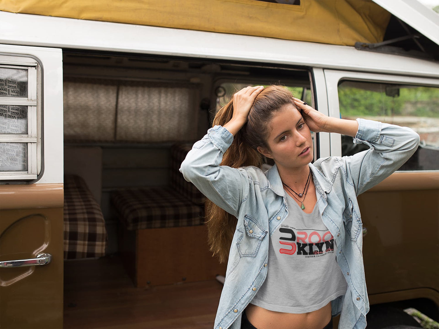 Woman standing in a van with hands on head, wearing a denim jacket and graphic t-shirt.