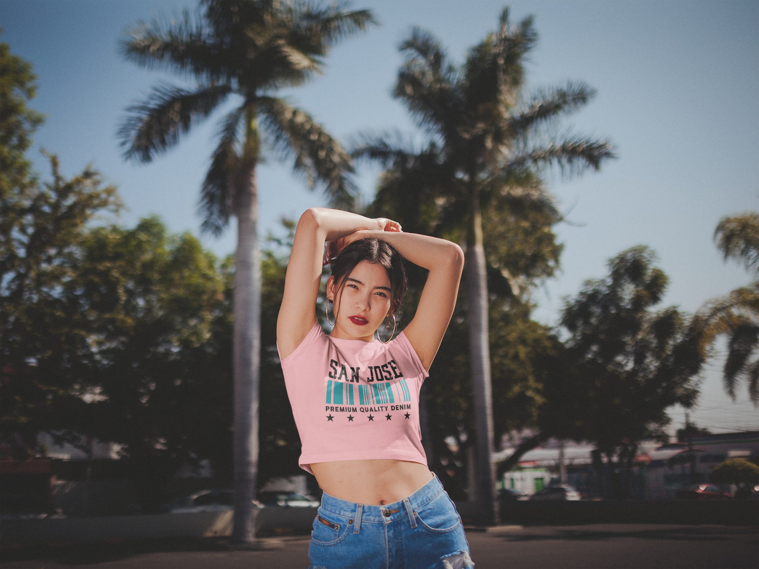 Woman wearing a pink crop top with text and blue jeans standing outdoors with palm trees in the background
