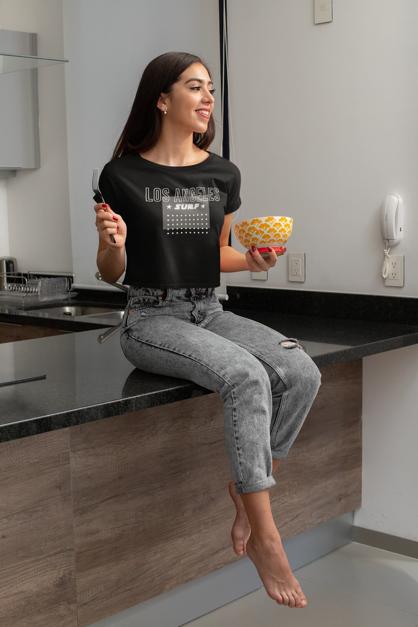 Woman sitting on a kitchen counter holding a bowl and a fork, wearing a black t-shirt with text.