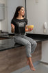 Woman sitting on a kitchen counter holding a bowl and a fork, wearing a black t-shirt with text.