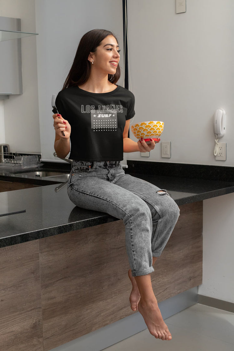 Woman sitting on a kitchen counter holding a bowl and a fork, wearing a black t-shirt with text.