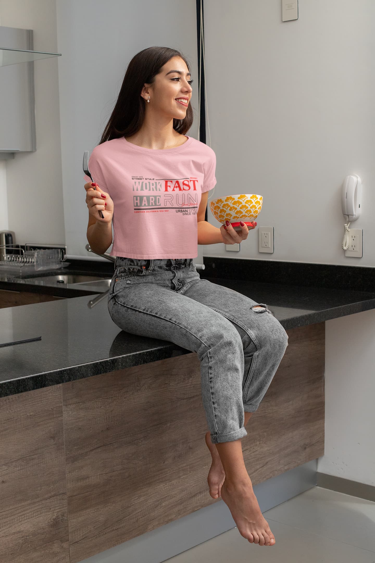 Woman sitting on a kitchen counter holding a bowl of fruit, wearing a pink t-shirt with text.