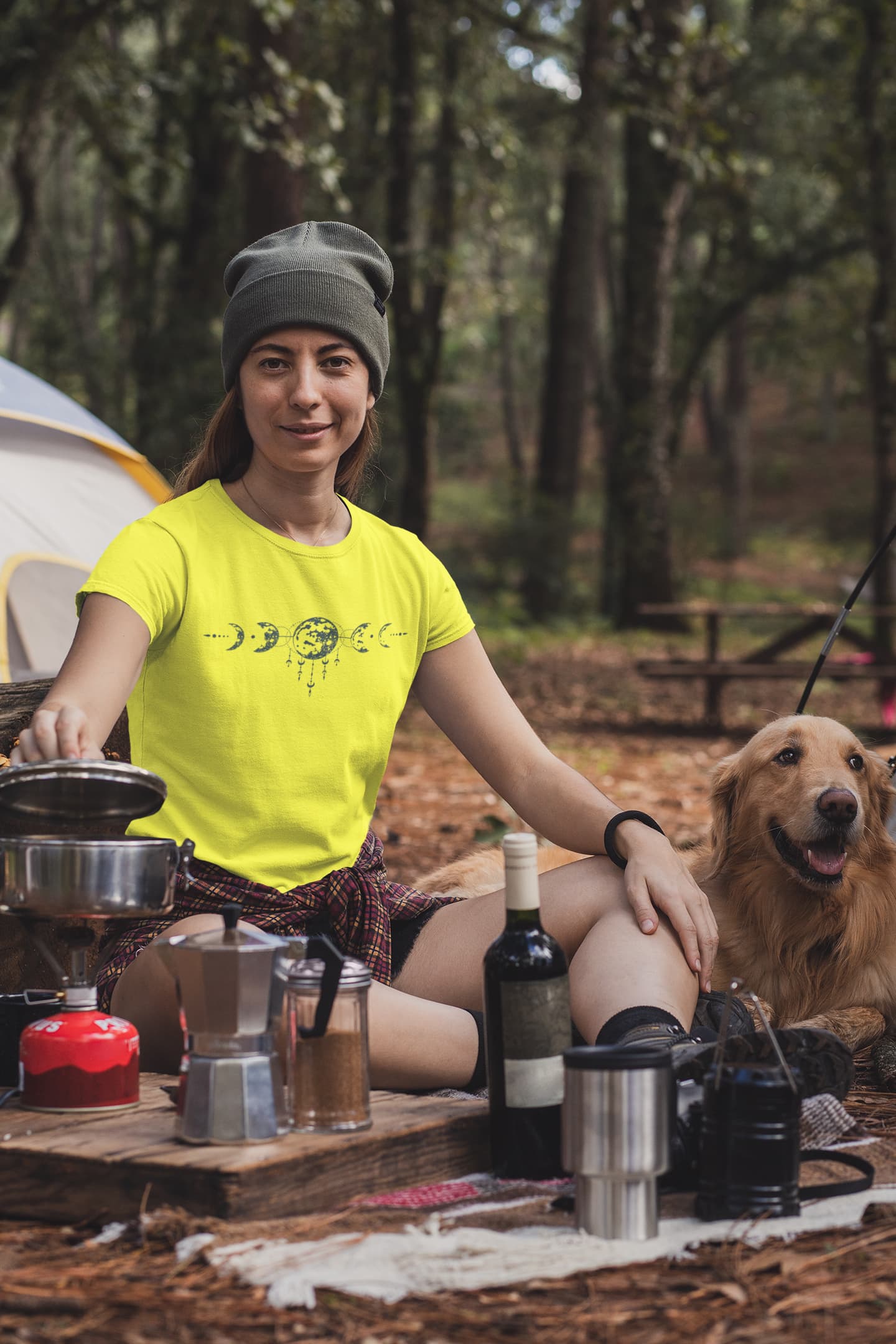 Woman in yellow shirt and gray beanie sitting outdoors with a dog, surrounded by camping gear.