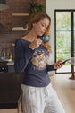 Woman in a kitchen holding a mug and phone, wearing a graphic t-shirt.