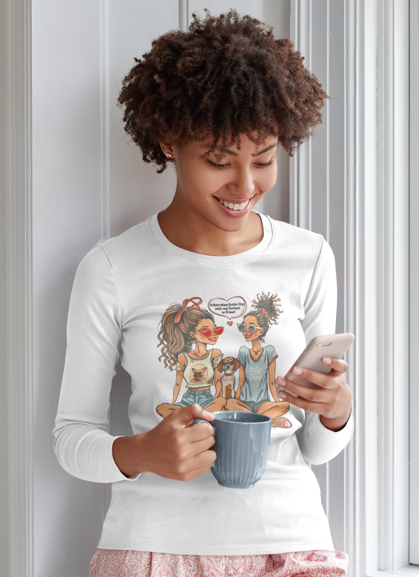 Woman wearing a white long-sleeve shirt with a graphic design, holding a mug and phone, against a light-colored wall.