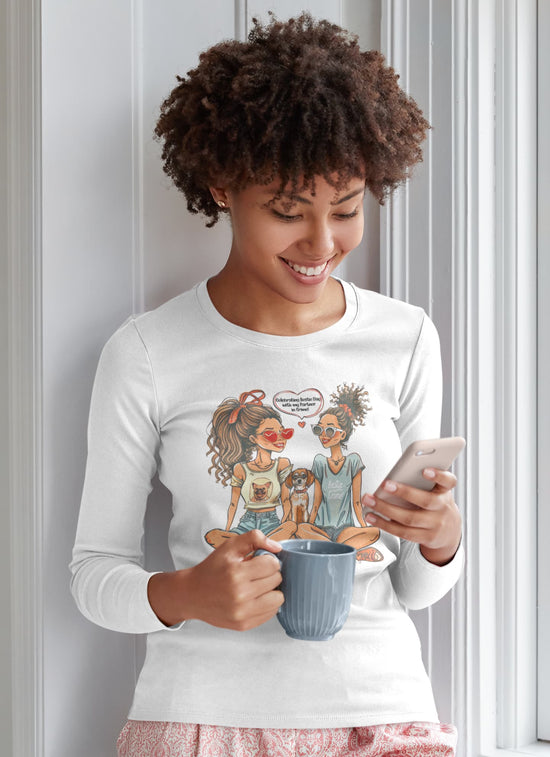 Woman wearing a white long-sleeve shirt with a graphic design, holding a mug and phone, against a light-colored wall.