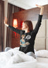 Woman in a black sweatshirt with a graphic design sitting on a bed in a room with a lamp and curtains.