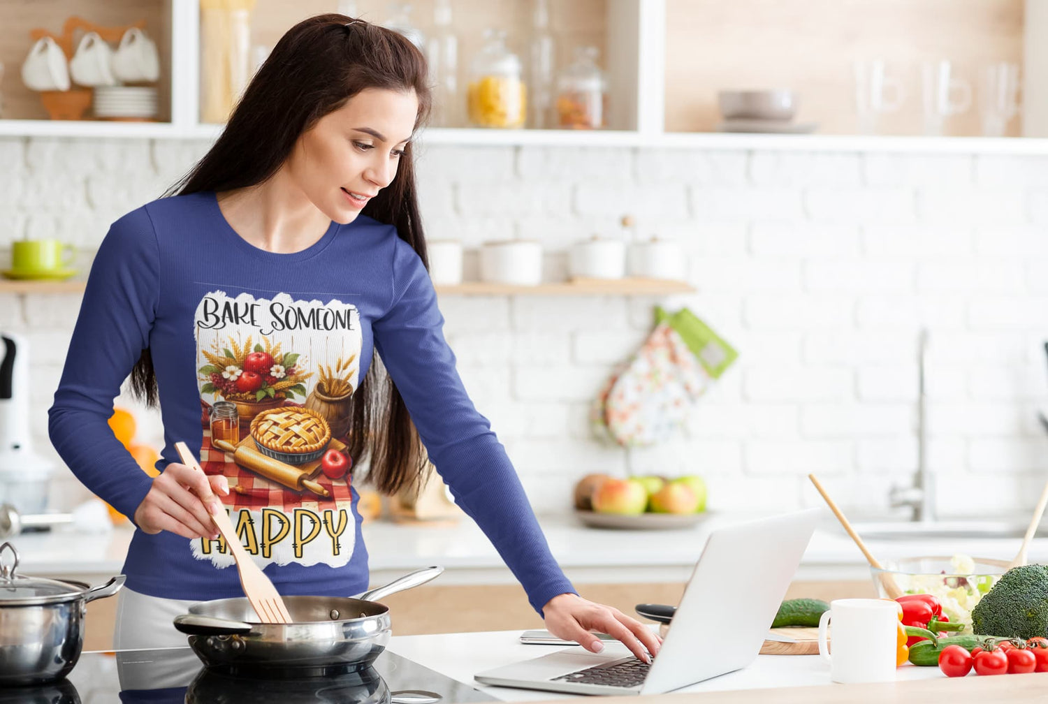 Woman in a kitchen wearing a blue shirt with a pizza design, using a laptop.
